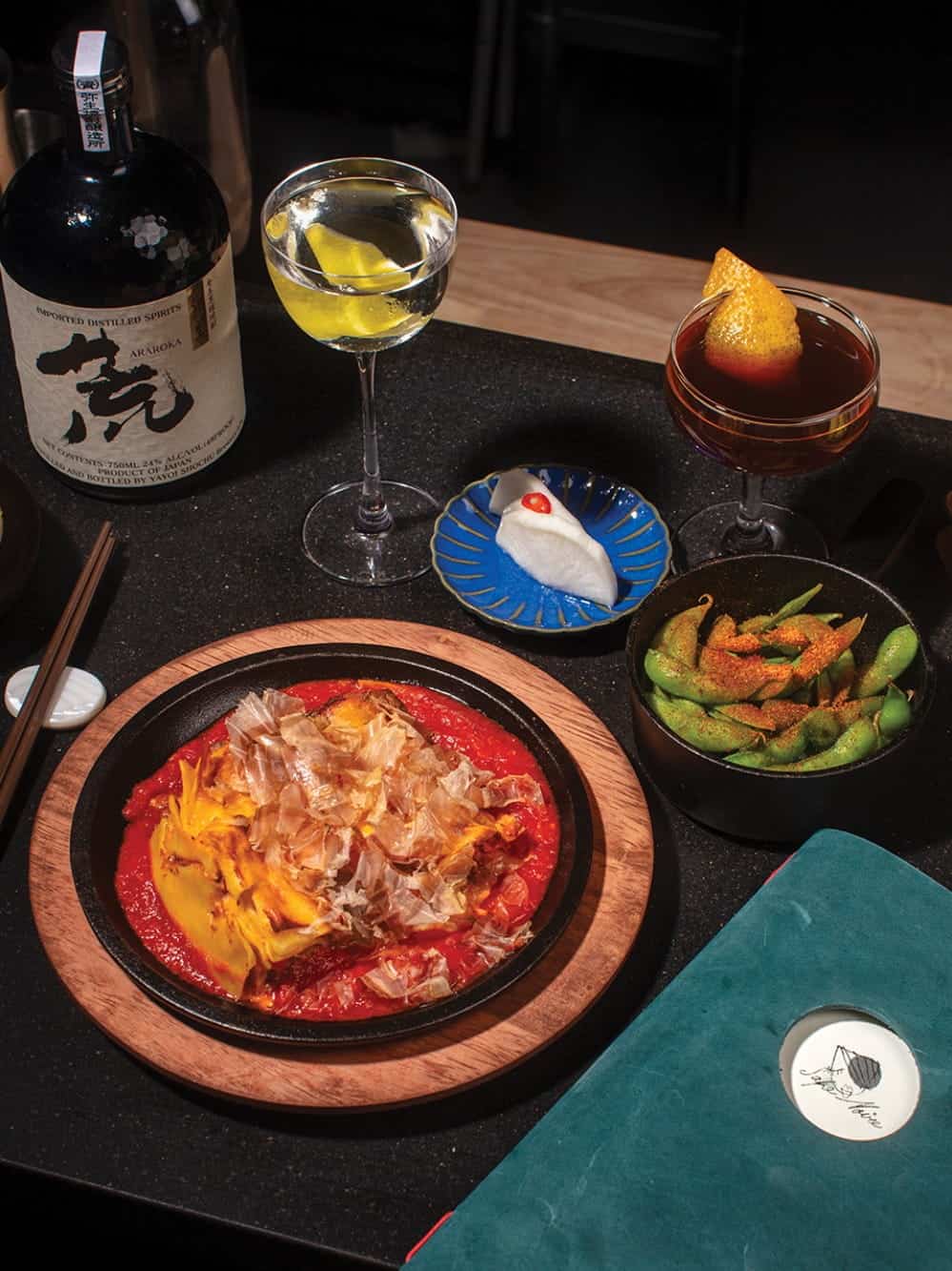 A tabletop at Louie’s Deli—one of the standout Seattle new restaurants—features a bottle of sake, two cocktails, a plate of edamame, a fish-shaped dessert, and a sizzling dish topped with bonito flakes on tomato sauce.