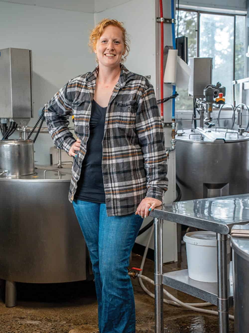 A person stands in a plaid shirt and jeans inside a modern industrial facility with stainless steel equipment and large containers, evoking the atmosphere of local creameries often featured on a Western Washington cheese road trip.