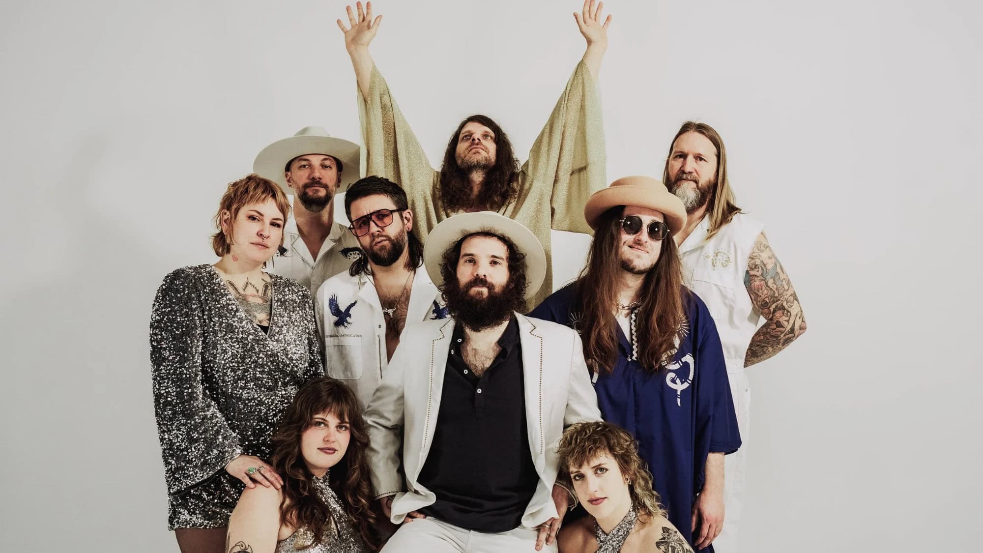 Nine adults pose against a plain white background, wearing various hats and eclectic outfits. One person in the back stands with arms raised. Most have long hair and some have visible tattoos.