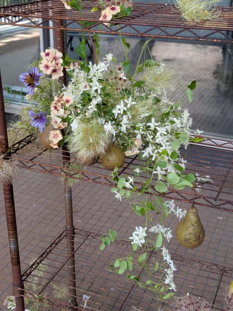 Metal shelves decorated with white and purple flowers, greenery, and two pears hanging among the floral arrangement.