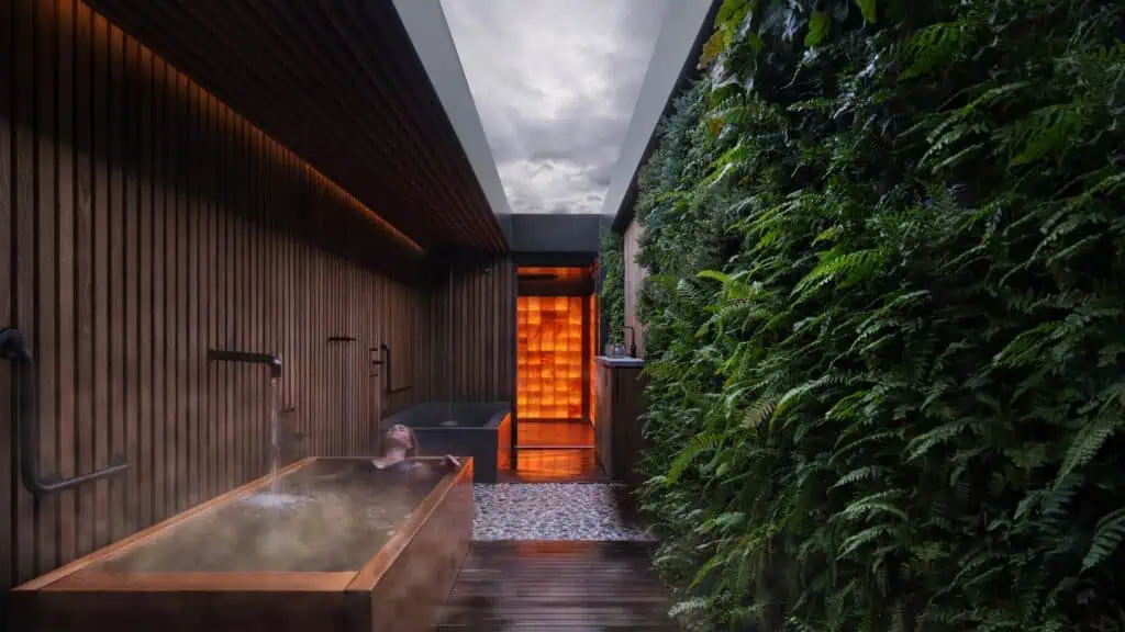 A person relaxes in a steaming wooden hot tub next to a lush vertical garden under a skylight, with wooden walls and an illuminated doorway in the background.