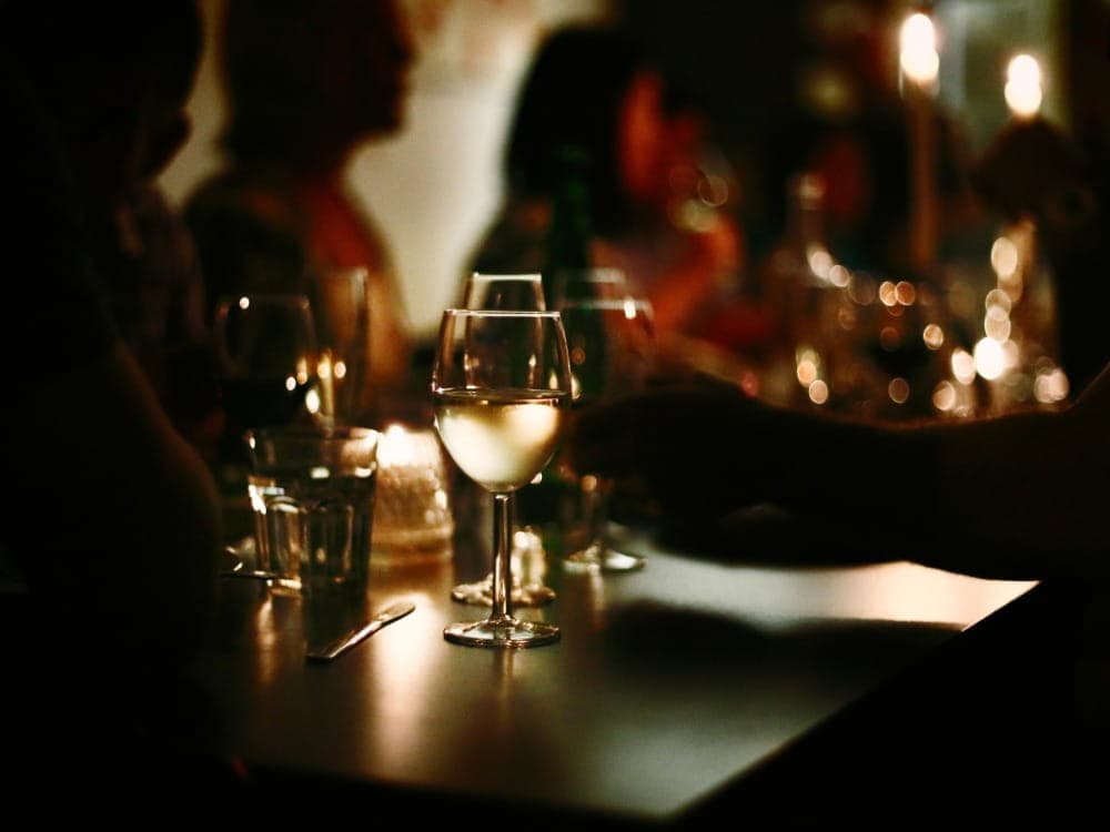 A dimly lit table with wine glasses, water glasses, and candles sets the scene; people are seated and conversing in the background, perhaps toasting to milestones like marriage.