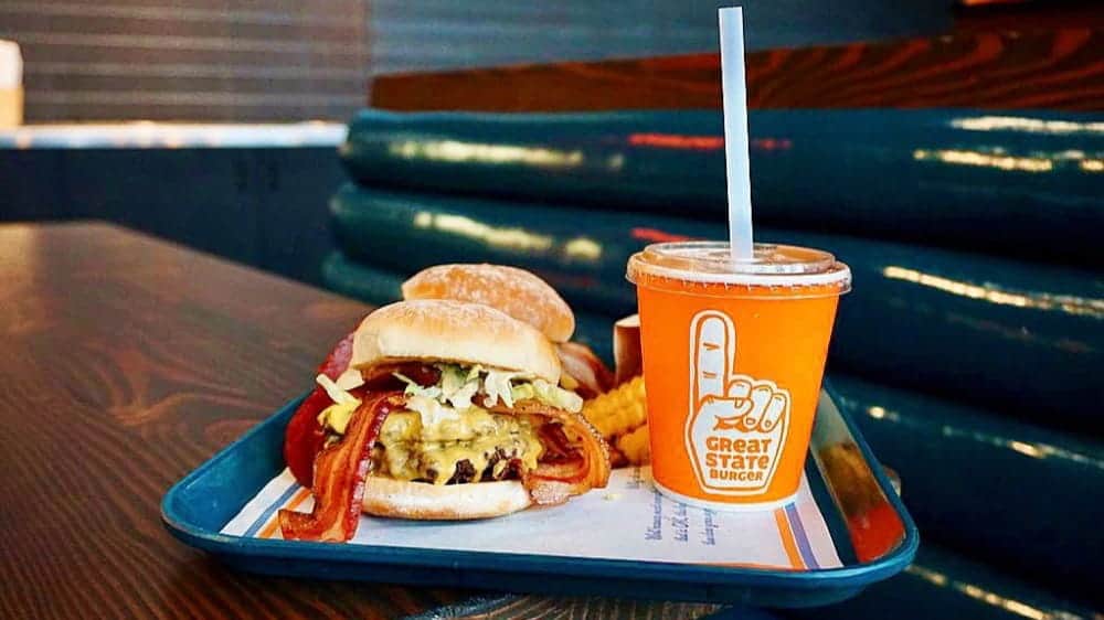 A tray with a bacon cheeseburger, crinkle-cut fries, and an orange cup with a straw sits invitingly on a wooden table in a restaurant—a classic Burger experience that’s perfect for sharing and celebrating any relationship.