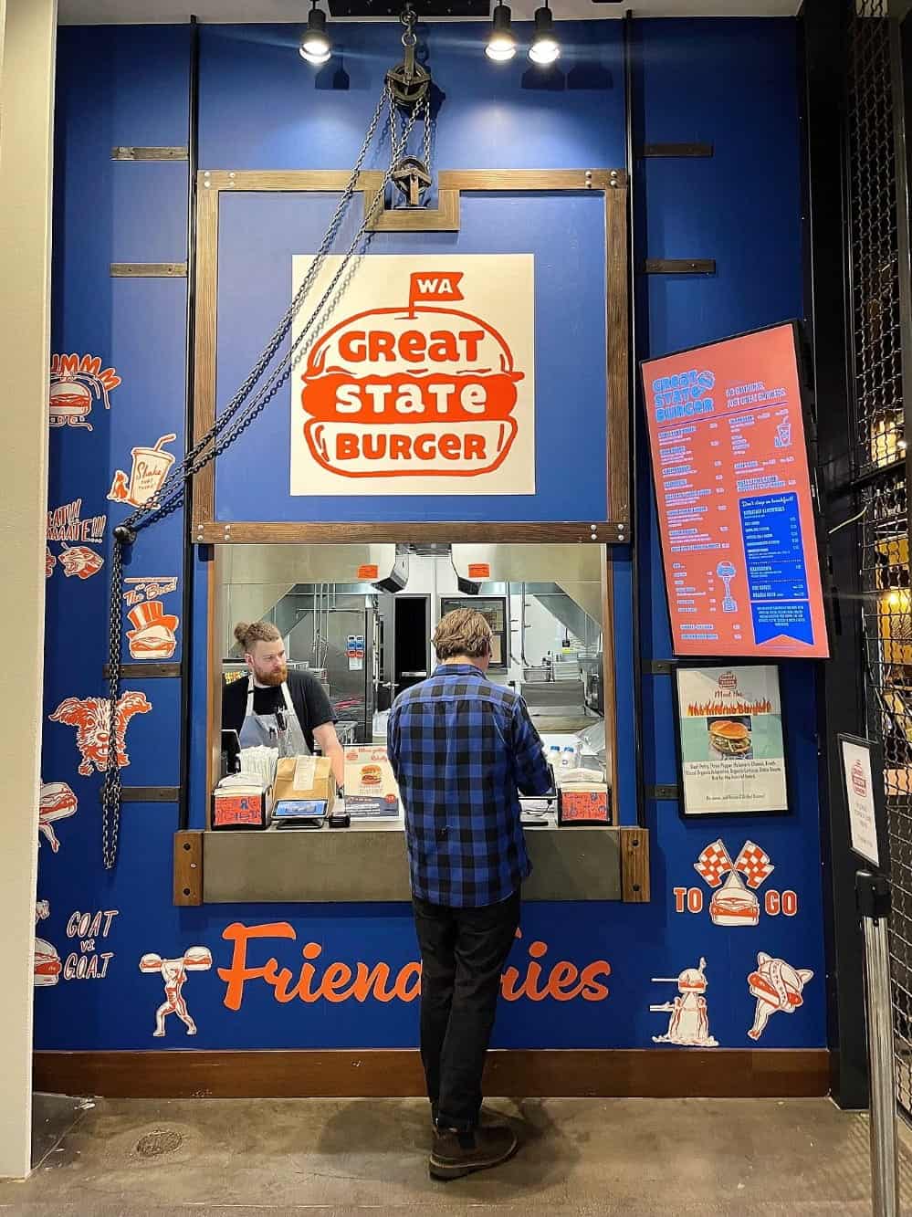 A person in a blue plaid shirt orders a burger at a Great State Burger counter with staff inside, surrounded by colorful signage and menu boards—perhaps savoring the kind of meal that almost saved my marriage.