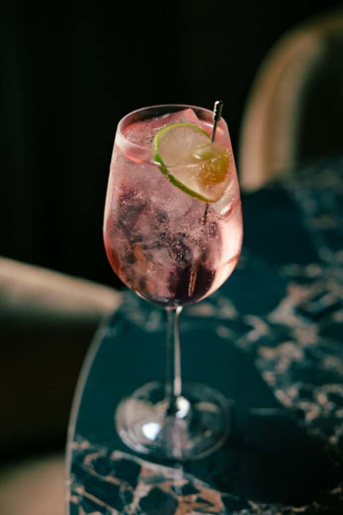 A stemmed glass with a pink sparkling drink, ice cubes, and a lime wheel garnish sits on a dark marble table.