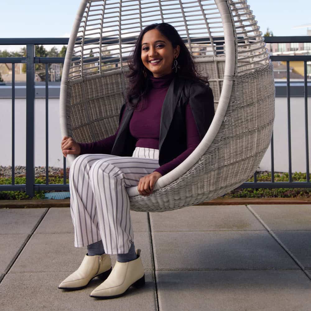 A woman in a striped outfit and cream boots sits in a hanging chair on a patio, smiling at the camera.