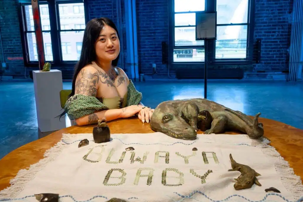 A woman with tattoos sits at a table displaying a sculpted animal and a cloth reading “GUWAYN BADI” in green letters, with small animal sculptures placed around the display—part of the Studio Sessions featuring artist Heidi Grace Acuña.