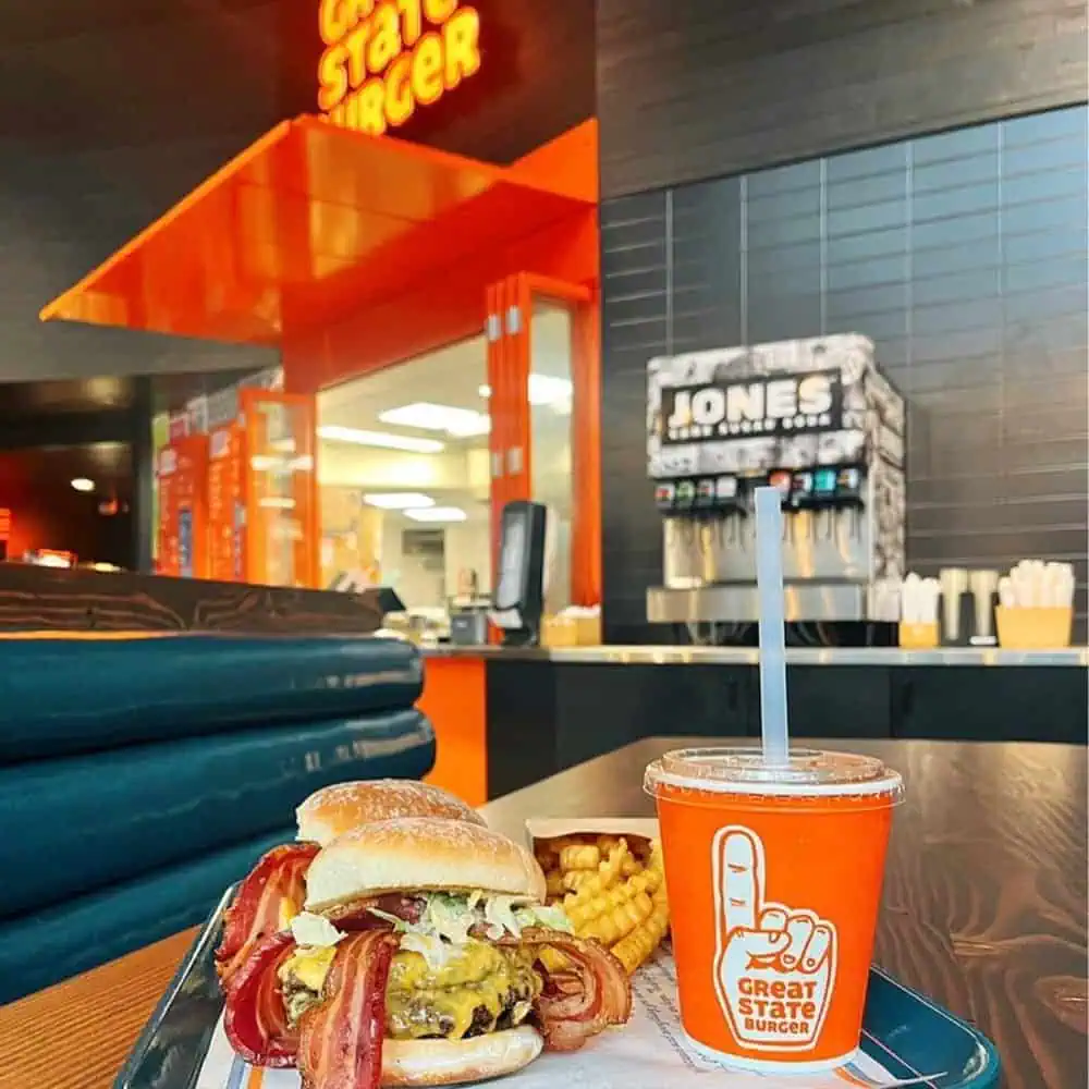 A table with a bacon cheeseburger, crinkle-cut fries, and a drink in an orange cup inside a brightly lit Great State Burger restaurant in Seattle—where you can enjoy delicious food crafted with care and fair wages for every team member.