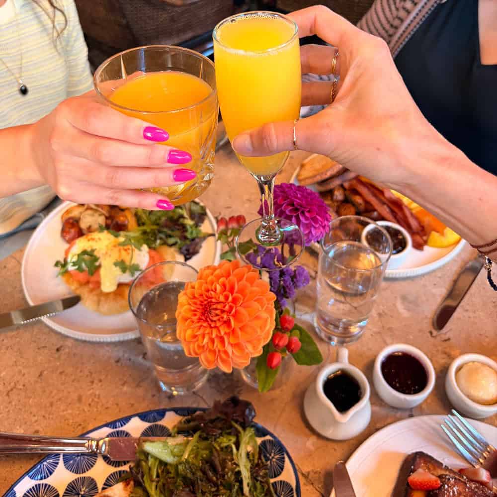 Two people toast with orange drinks over a brunch table set with plates of food, water glasses, flowers, and small bowls of condiments—perhaps at the latest spot featured in Restaurant Roundup.