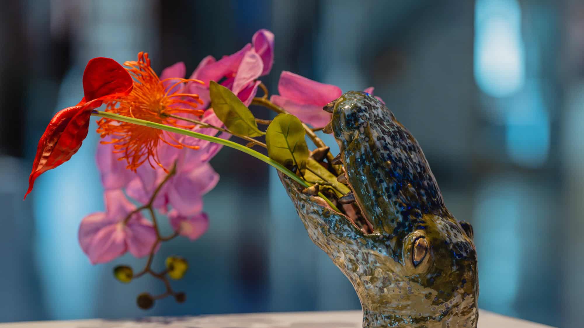 A ceramic sculpture of an open-mouthed crocodile, inspired by Crocodile Guardians, holds colorful flowers and green leaves against a blurred background.