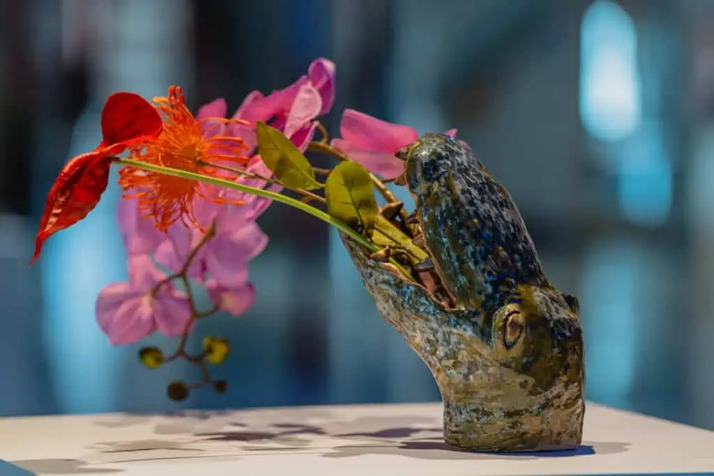 A ceramic sculpture of an animal head, possibly a crocodile, holds an arrangement of colorful flowers and greenery against a blurred background.