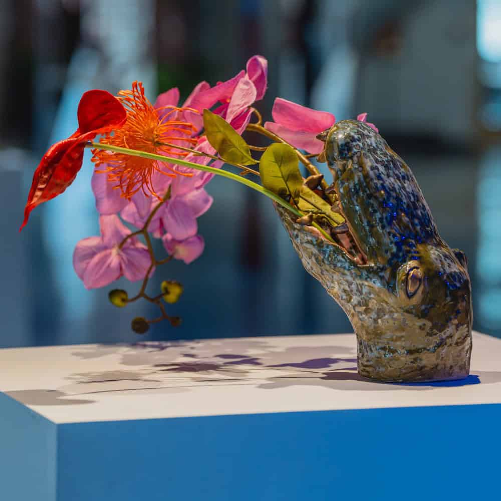 Ceramic vase shaped like a lizard head, inspired by Crocodile Guardians, holds a colorful arrangement of red, orange, and pink flowers, displayed on a white pedestal.