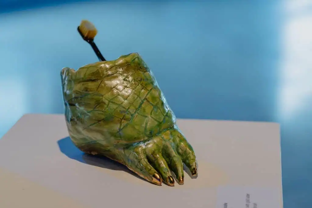 A green ceramic sculpture shaped like a reptilian foot holds a toothbrush, displayed on a white surface with a blurred blue background.