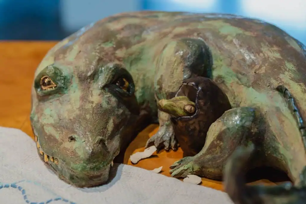 A green, lizard-like sculpture with a baby hatching from an egg beside it, both posed on a wooden surface with part of a white cloth visible in the foreground.