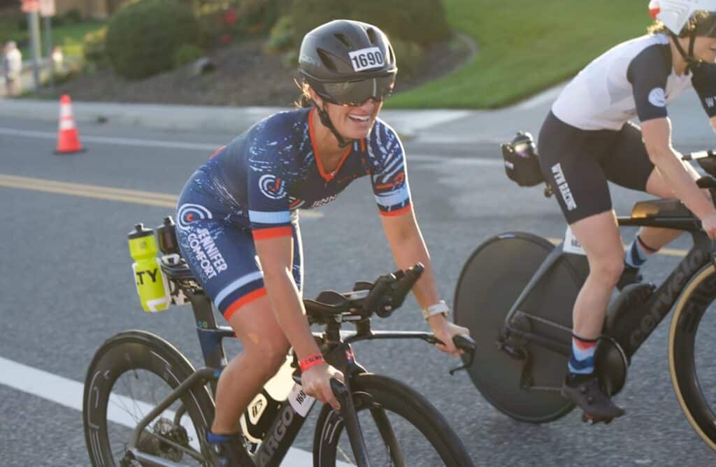 Two cyclists in racing gear ride on a sunlit road, showcasing their fortitude; the cyclist in front smiles while wearing a helmet labeled “1690,” embodying the determination often seen in triathlon events.
