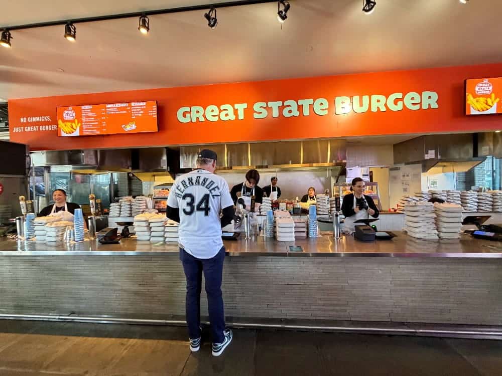 A customer stands at the counter of Great State Burger in Seattle, where staff prepare and serve delicious grass-fed beef burgers with stacks of takeout boxes visible.