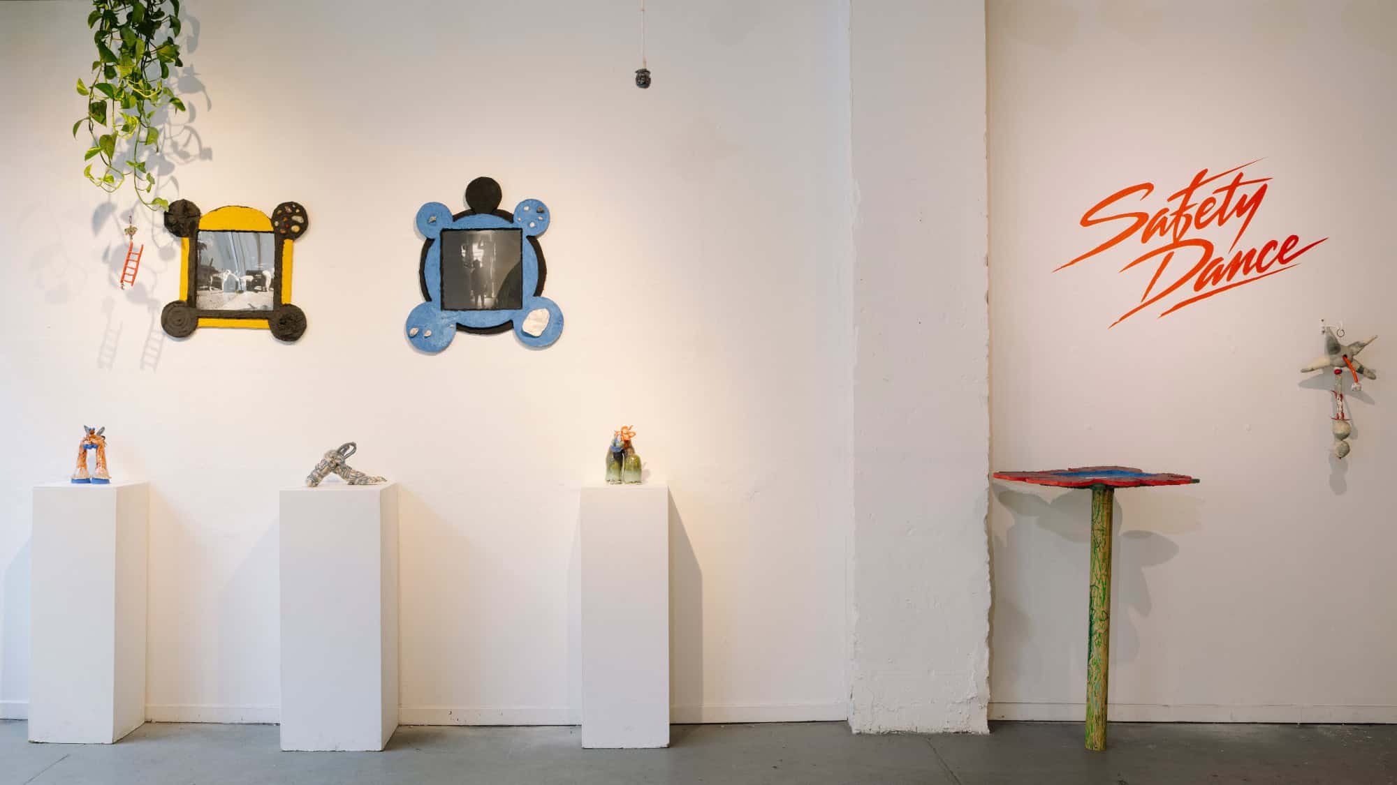 Art gallery wall with framed photos, small sculptures on pedestals, a “Safety Dance” text mural, and a plant hanging from the ceiling.