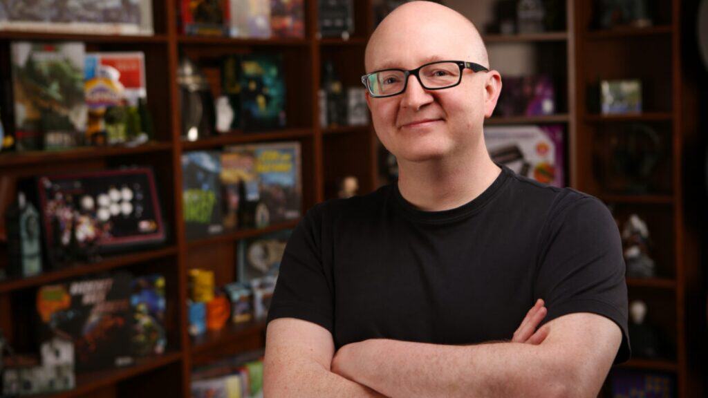 A bald person wearing glasses and a black t-shirt stands with arms crossed in front of shelves filled with books, games, and Magic collectibles—looking stronger than ever, ready for the next PAX.