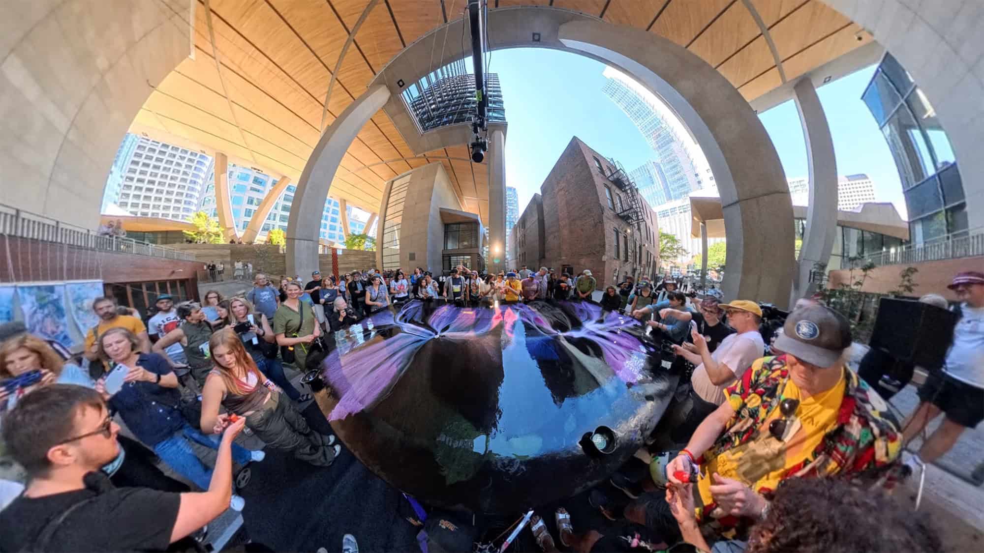 A crowd gathers around a large reflective sculpture under an outdoor architectural structure in an urban setting during daylight.