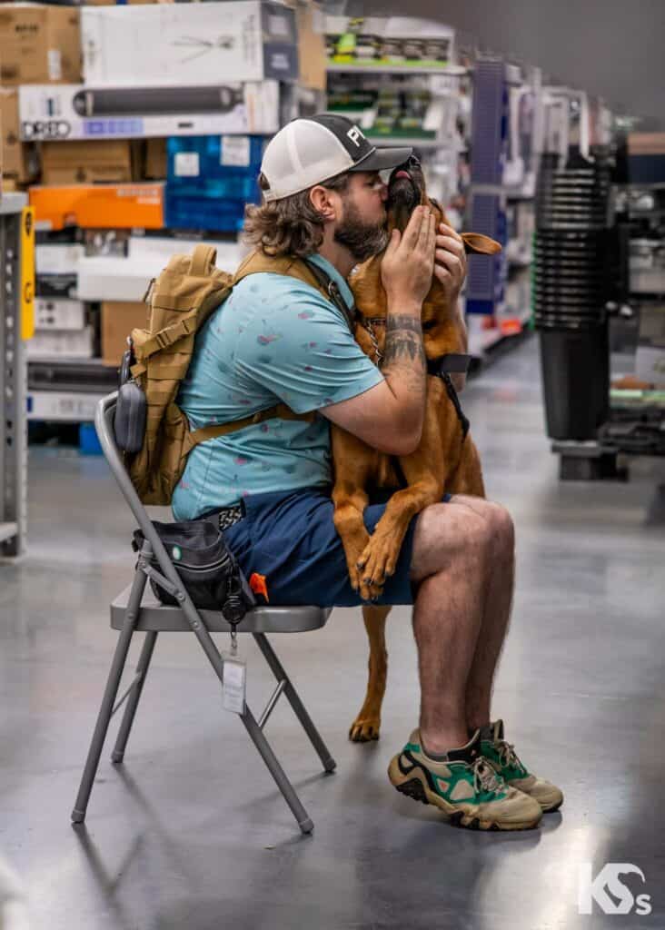 A man sits on a folding chair in a store aisle, embracing and kissing a brown dog. The man wears a cap, shorts, and sandals, and has a tan backpack. The dog has its front paws on his lap.