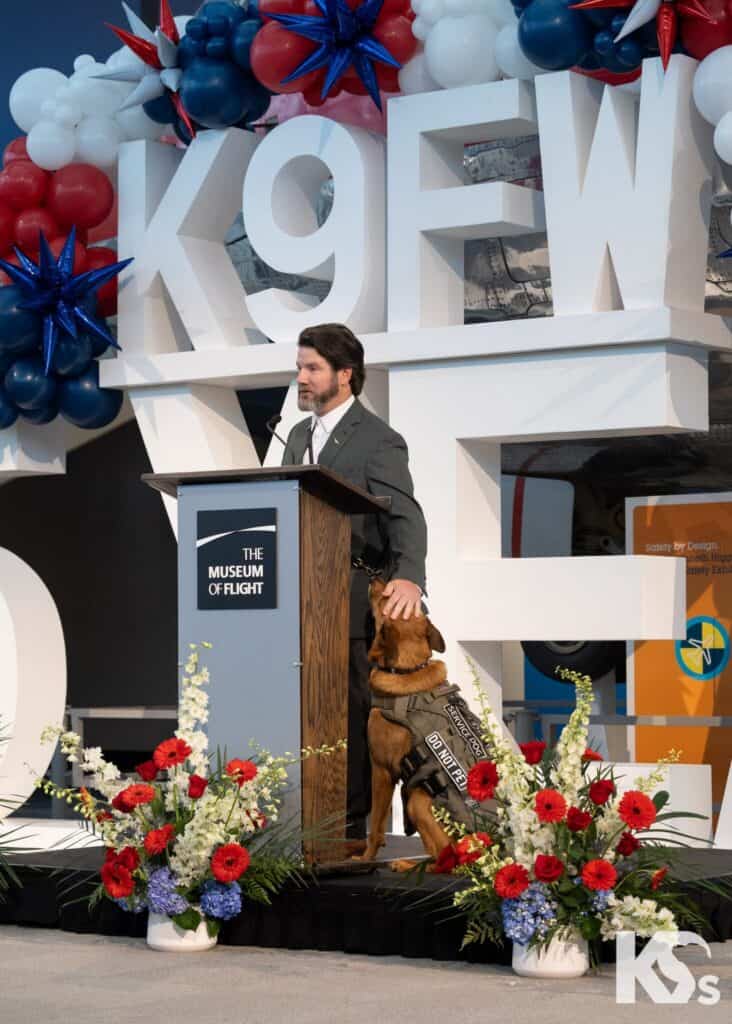 A man in a suit stands at a podium labeled “The Museum of Flight” with a service dog by his side, surrounded by flowers and large balloon letters reading “K9FW.”.