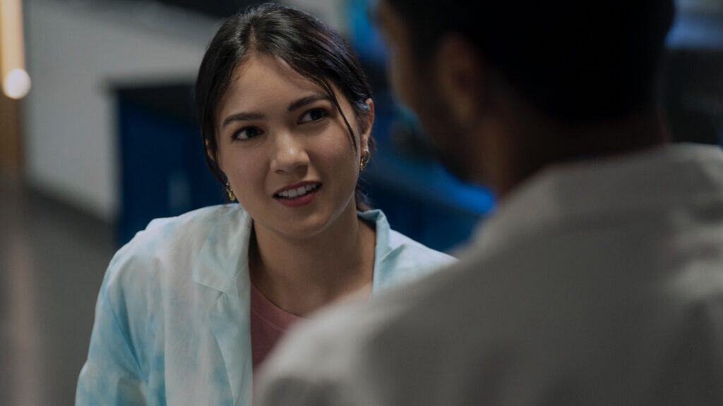 A woman in a lab coat looks at a man in conversation, with lab equipment and blue cabinets visible in the background.