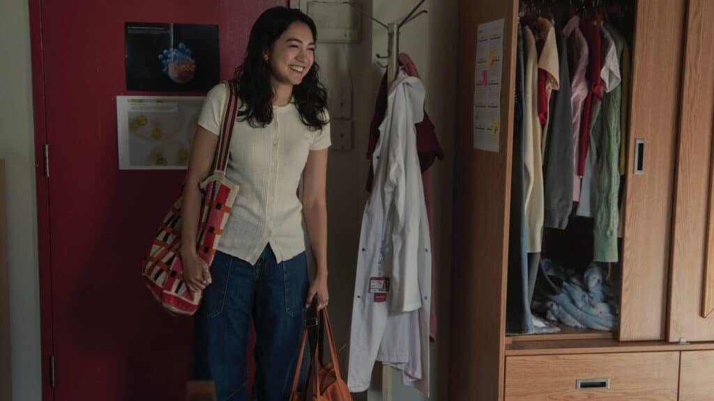 A woman stands smiling in a room with a red door, holding two bags. Clothes hang in an open wardrobe and a lab coat hangs on a hook.