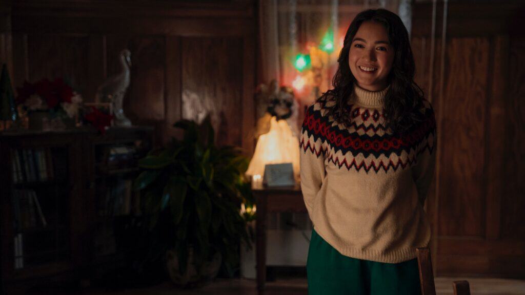 A woman in a patterned sweater stands indoors, smiling, with warm lighting and festive decorations in the background.