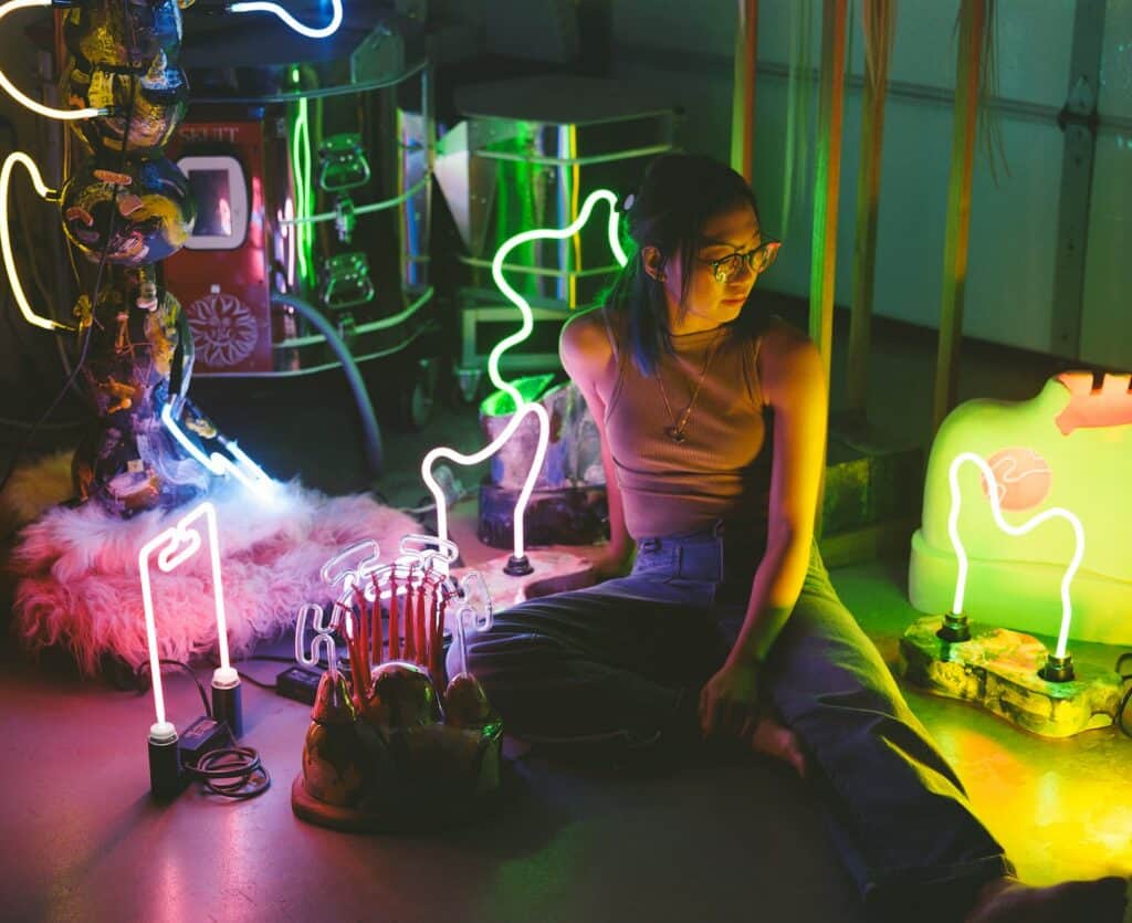 A person sits on the floor surrounded by colorful neon lights in various shapes, creating a vibrant and artistic atmosphere.