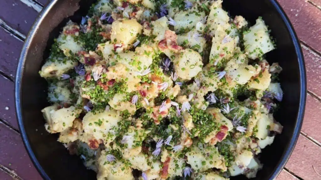 A bowl of potato salad with chopped herbs, crumbled bacon, and small purple edible flowers on top.