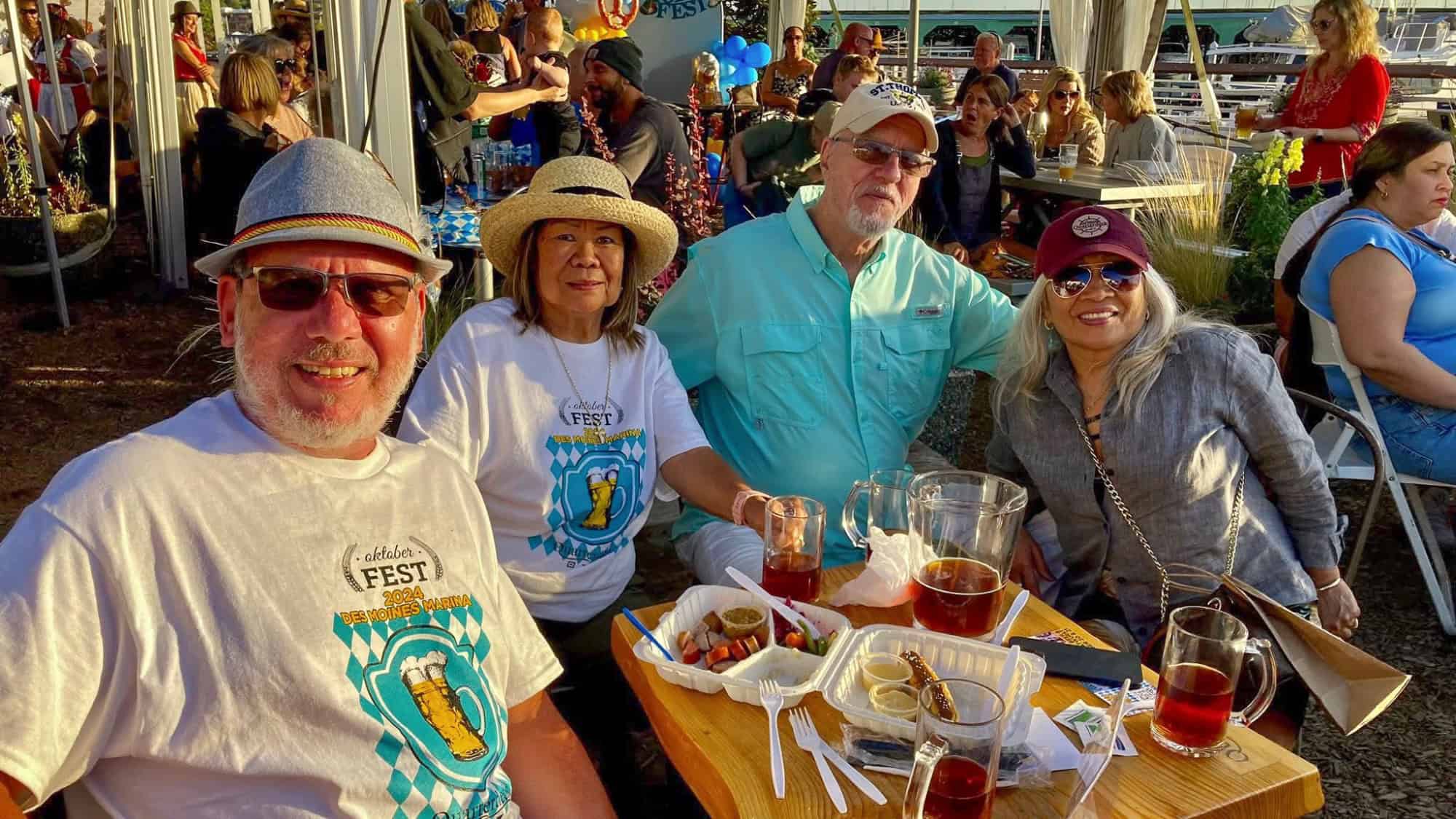 Four adults sit at an outdoor table with drinks and food trays, wearing hats and sunglasses, during a festival with other people in the background.