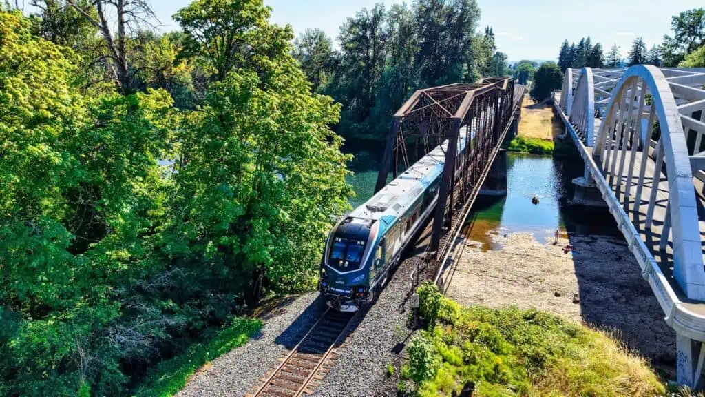A passenger train crosses a metal railway bridge over a river, surrounded by green trees and a pedestrian bridge to the right.