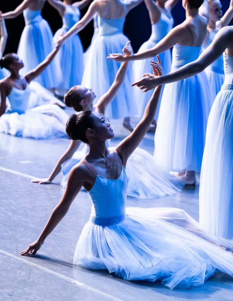 Ballet dancers in white tutus perform on stage, with one dancer in the foreground kneeling and reaching upward.
