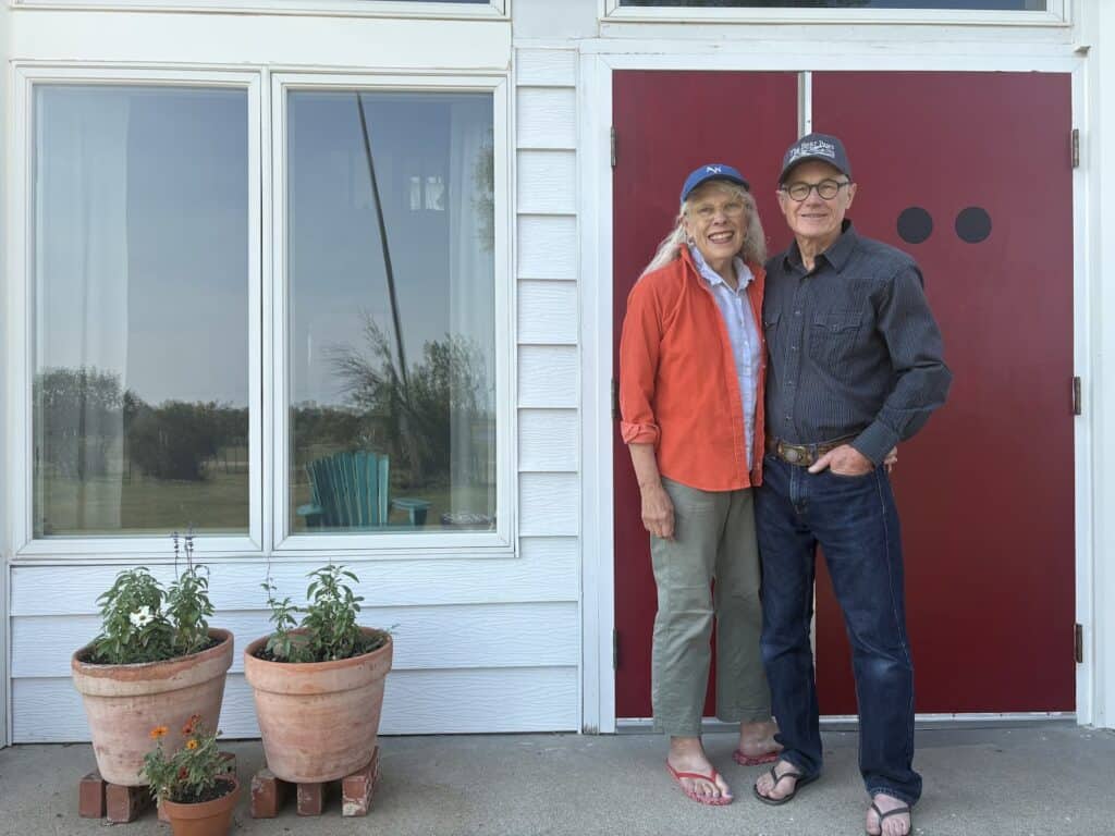 Two older adults stand smiling in front of a red door and white house, with three potted plants on the ground nearby and a window reflecting an outdoor scene.