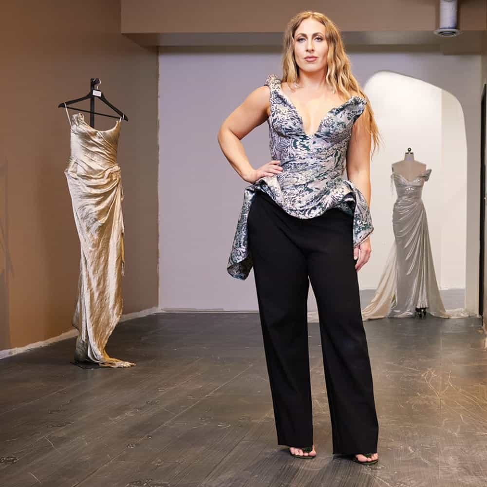 Woman stands in a room wearing a patterned top and black pants; two metallic gowns, inspired by Seattle public art, on display in the background.