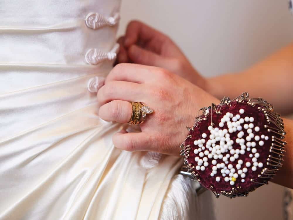 A person adjusts the back of a white satin dress, wearing a pin cushion bracelet, inspired by the intricate designs of Seattle manhole covers.