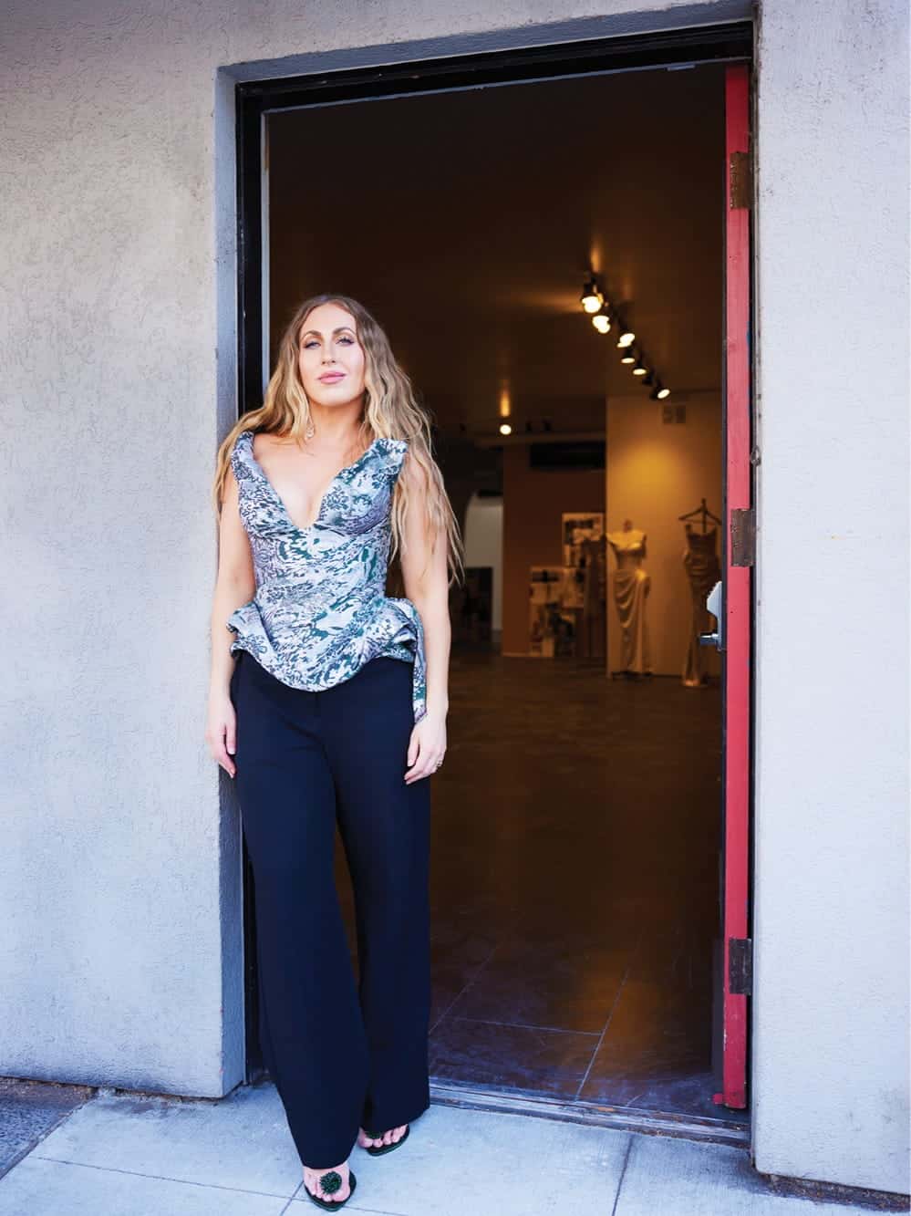 Woman with long, wavy hair stands in a doorway wearing a silver patterned top and black pants, echoing the creative spirit of Seattle public art. Dimly lit room with dresses behind her.