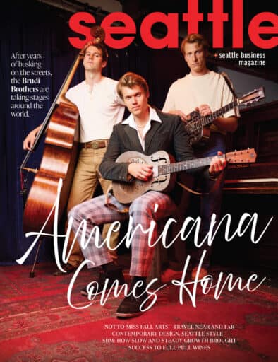 Three men with musical instruments pose onstage in front of a piano and upright bass, featured on the cover of Seattle magazine with the headline “Americana Comes Home.”.