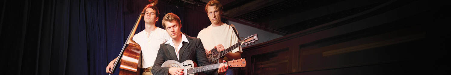 Three men pose on stage with musical instruments: one with a double bass, one seated with a guitar, and one standing with an electric guitar; dark curtains and dim lighting in the background.