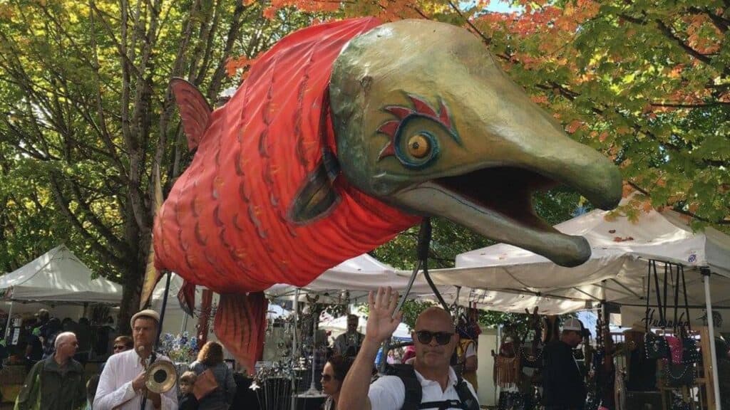 A large fish puppet is carried by people at an outdoor event with tents and trees in the background.
