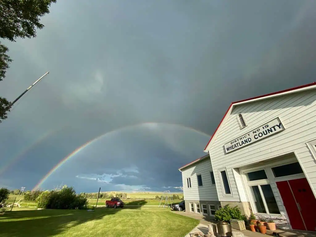 A double rainbow arcs over a grassy yard beside a white building labeled “District No. 15 Wheatland County” under a partly cloudy sky.