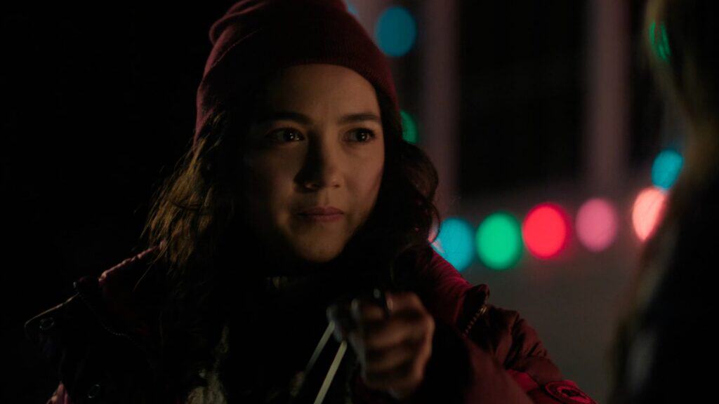 A young person in a red hat and jacket stands outdoors at night, holding an object forward, as colorful blurred lights glow—just like a scene from a good joke by Rachel Marsh.