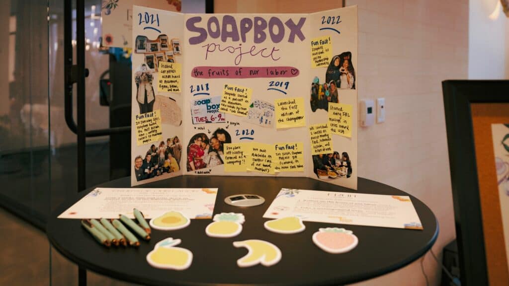 A trifold display titled “Soapbox Project” stands on a round table with papers, stickers of fruit shapes, and pens arranged in front of it.