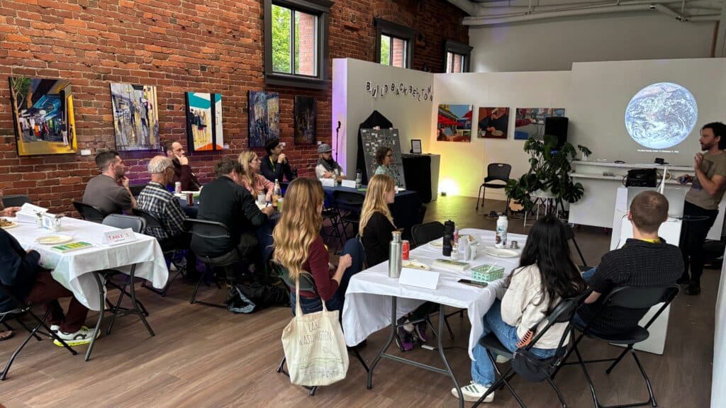 A group of people sit at tables listening to a speaker in a brick-walled room with art on the walls and a projected image of Earth.