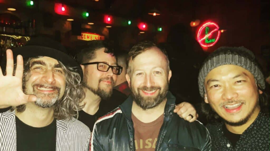 Four men pose and smile in a dimly lit bar with colorful lights and a neon sign in the background, capturing the lively atmosphere of a Stereo Embers live concert.
