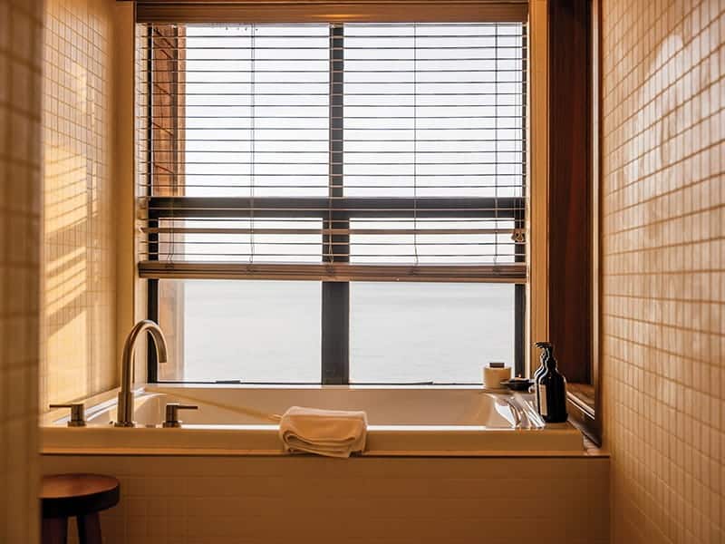 A bathtub set against a large window with horizontal blinds overlooking the water at the Inn at Langley.