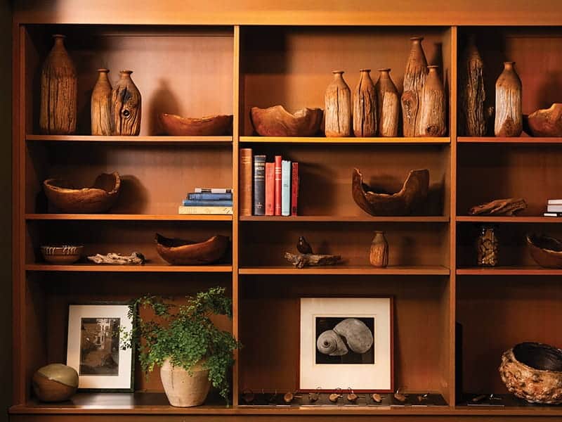 Wood shelving unit at Passage Restaurant displaying wooden vases, carved bowls, books, a potted plant, and framed artwork.