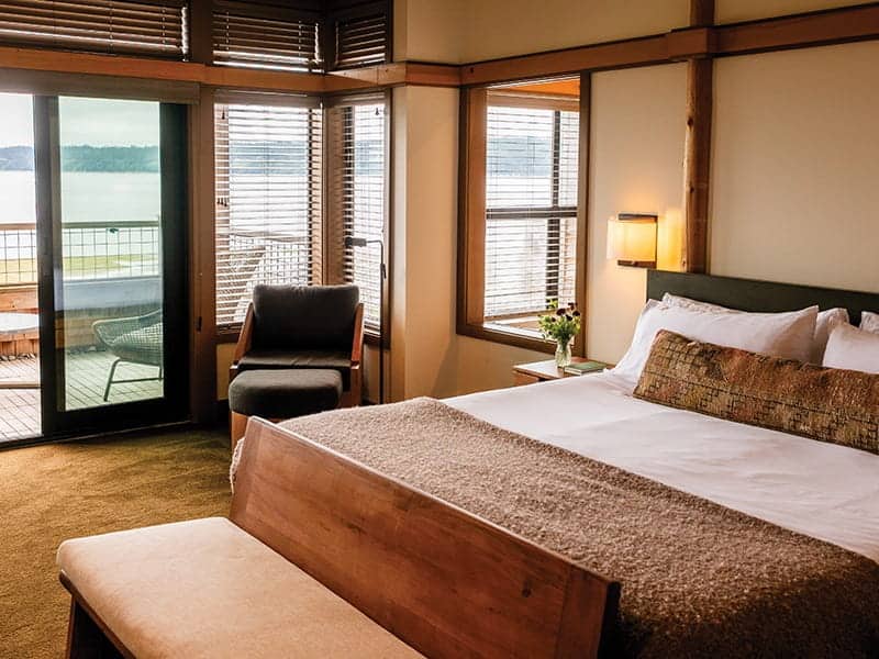 A queen bedroom at a Langley, WA hotel, featuring a lounge chair with an ottoman and a sliding glass door opening to a balcony with a water view.