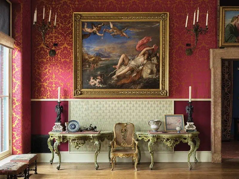 A painting of the Abduction of Europa in a gold frame, centered above console tables and an armchair against red damask wallpaper, typical of top art collections.