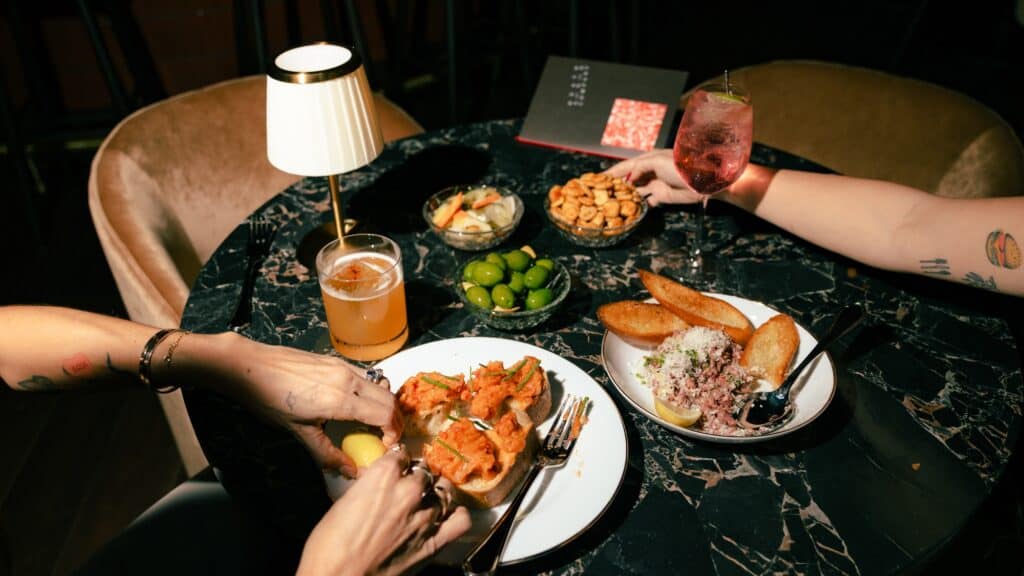 Two people sit at a black marble table in Death & Co. Pioneer Square, enjoying cocktails, olives, nuts, bruschetta, and a meat dish as they reach for food under a small lamp.